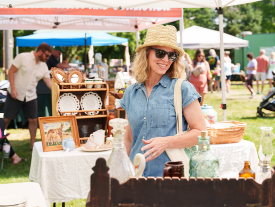 an older woman with shoulder length, blonde curly hair wearing a denim shirt, straw hat and sun glasses shopping at an open market